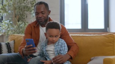 Evde tuhaf bir eğlence. Oyuncu Afrikalı Amerikalı adam oğluyla vakit geçiriyor, sosyal medya için akıllı telefondan özçekim yapıyor, kameraya yüzünü ekşitiyor, görüntüyü takip ediyor, yavaş çekim yapıyor.