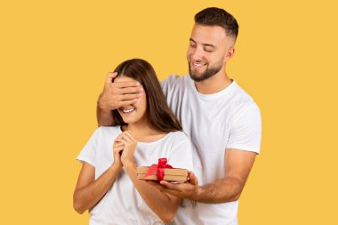 Neşeli genç Avrupalı adam kadınlara gözlerini kapattı, sarı stüdyo arka planında izole edilmiş bir kutu hediye etti. Çiftler tatilleri, doğum günlerini ve yeni yılı, Noel hediyelerini, sevgililer günü sürprizlerini severler.