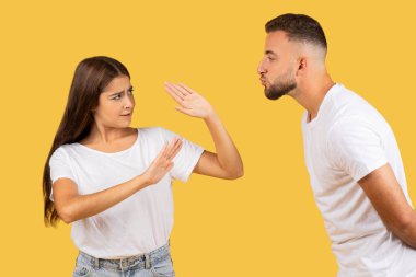 Beyaz tişörtlü kızgın Avrupalı kadın adamı durdurup, sarı arka planda izole bir şekilde öpüşmek istiyor. İlişki sorunları, taciz, anlaşmazlık veya kişisel sınırlarla mücadele