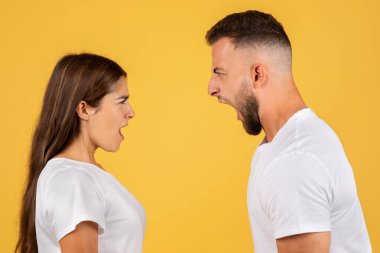 Beyaz tişörtlü üzgün Avrupalı çift çığlıklar atıyor. Ağızları açık, sarı arka planda izole edilmiş bir şekilde kavga ediyorlar. İlişki sorunları, skandal ve çatışma