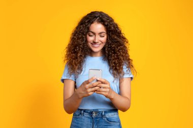 Kıvırcık saçlı, gülümseyen genç bir kadın akıllı telefonuyla mesajlaşmaktan zevk alıyor, güneşli sarı arka planda duruyor, rahat ve mutlu tavırlar sergiliyor.