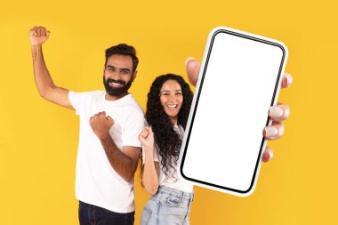 Mutlu Ortadoğu çifti, sarı arkaplan üzerinde boş ekranlı büyük bir akıllı telefon gösteriyor. İkisi de başarı için işaret ediyor. Teknoloji ve internet uygulamaları için ideal.
