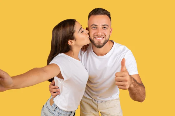 穿着白色T恤衫的积极的年轻高加索女人吻着被检查的男人 玩得开心 享受约会 展示拇指向上的标志 在电话上自拍 在黄色的工作室背景下被隔离 爱的认可 博客的照片 — 图库照片