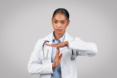 Beyaz önlüklü bin yıllık kendine güvenen siyahi kadın doktor dur işareti yapıyor, mola veriyor, gri arka planda izole edilmiş, stüdyo. Sağlık hizmeti jestin bitmesine yardım eder, sorunlarla mücadele eder