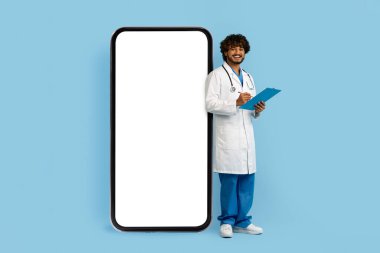 Medikal üniforma giyen neşeli, genç Hintli doktor terapist elinde pano beyaz boş ekranlı, mavi stüdyo arka planında izole edilmiş kocaman bir telefonun yanında duruyor.