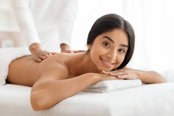 Young beautiful woman receiving relaxing back massage from professional therapist in spa salon, closeup shot of smiling attractive indian lady enjoying body treatment in beauty studio