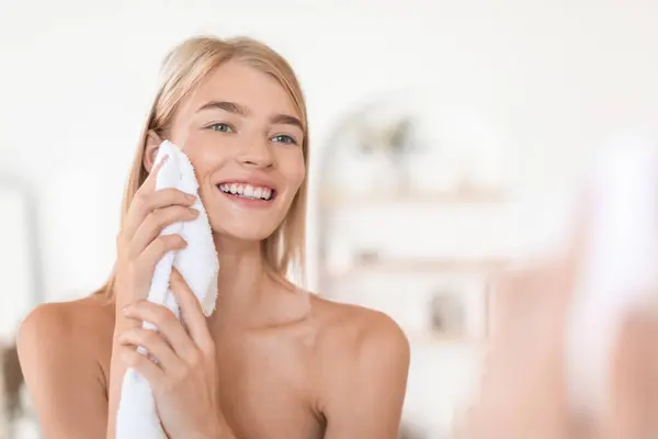 Blonde European woman gently dries face with white towel, smiling to her reflection in mirror, standing shirtless in modern bathroom at home. Facial skincare routine and beauty pampering
