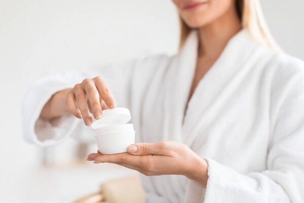 Cropped shot of lady holding and opening cosmetic jar with moisturizer cream, moisturizing skin and caring for herself in modern bathroom indoor. Beauty and wellness routine