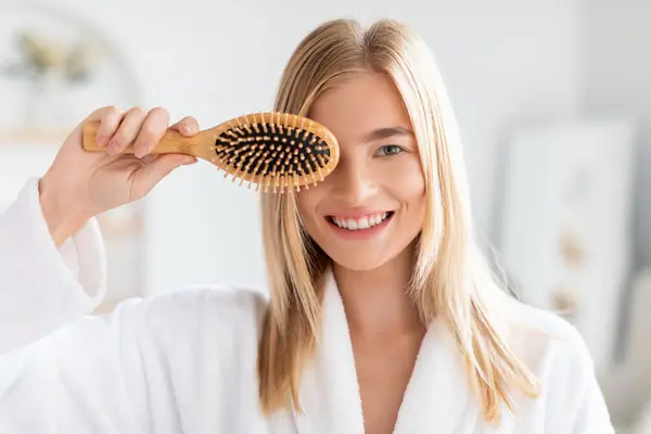 Blonde woman in white bathrobe playfully covers one eye with hairbrush, smiling to camera in home bathroom. Lady enjoys morning of self care, detangling her hair with wooden brush