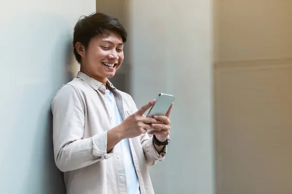 Positive young chinese guy employee have break, standing next to office building, using phone and smiling outdoors, copy space for business mobile application concept