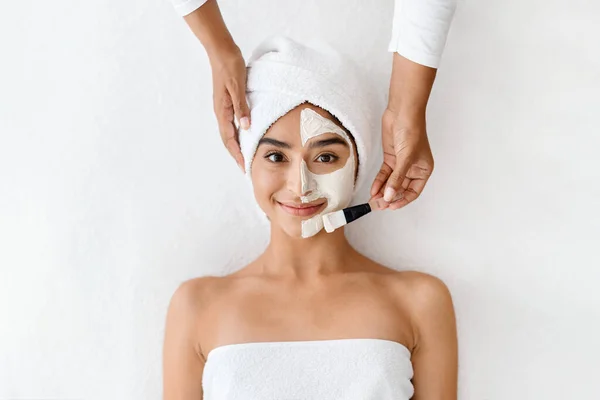 A beautiful Eastern woman enjoys a rejuvenating facial treatment at a luxury spa, where a professional skin therapist gently spreads a cleansing clay mask over half of her face. Before and after