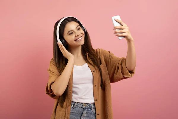 快乐的少女 戴着无线耳机 在智能手机上自拍 微笑的少女 在手机摄像机前摆姿势 在工作室里站在粉色背景上开心 自由自在 — 图库照片