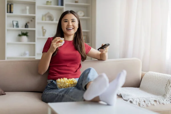 Happy relaxed asian woman watching tv and eating popcorn at home, smiling female switching channels with remote controller, resting on couch in cozy living room, enjoying domestic weekend, copy space