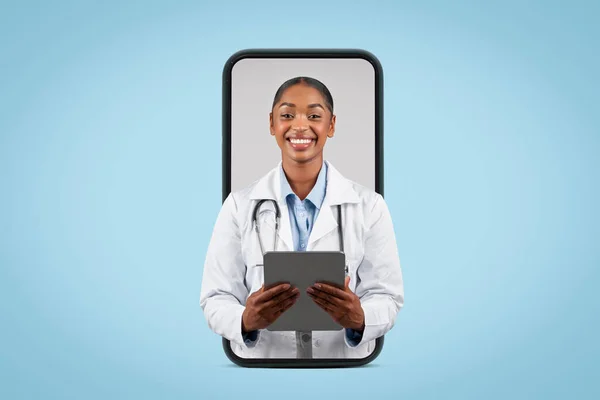 Positive young black lady doctor therapist in white coat typing on tablet, on big phone screen, isolated on blue background, studio. Recommend medical health care app, consultation online