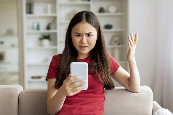 不满意的年轻亚洲女人坐在沙发上看着智能手机 焦虑的韩国女人在手机上阅读讯息 对坏消息做出情绪反应 复制空间 — 图库照片