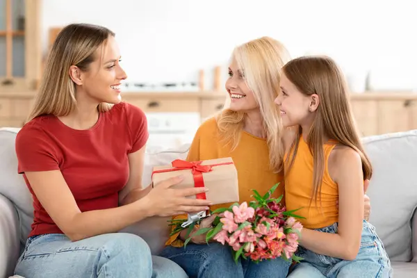 Three generations of women on Mothers Day at home. A cheerful family moment as a grandmother is greeted with International Womens Day gifts by her daughter and granddaughter