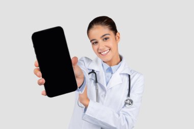 Beyaz önlüklü gülümseyen Kafkas kadın doktor beyaz perdeli akıllı telefon, gri stüdyo arka planında izole. Web sitesi tavsiyesi, tıbbi sağlık uygulaması