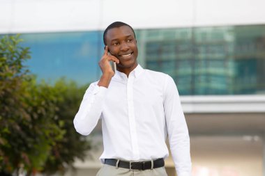 İletişim Konsepti. Dışarıda cep telefonuyla konuşan yakışıklı siyahi iş adamı, Beyaz gömlekli gülümseyen Afrikalı Amerikalı genç adam dışarıda yürürken keyifli cep telefonu sohbetleri yapıyor.