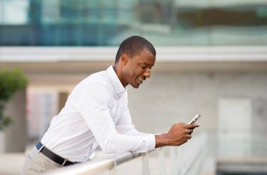 Gülümseyen Siyah İşadamı Demiryolu 'na yaslanıyor ve akıllı telefonu açık havada kullanıyor, genç Afrikalı Amerikalı erkek girişimci dışarıda cep telefonunu kullanıyor, cep telefonunda mesajlaşıyor,