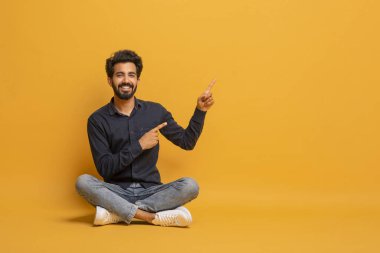 Neşeli Hintli adam yerde oturup fotokopi alanını işaret ediyor, mutlu gülümseyen doğu çocuğu iyi bir fikir ya da çözüm öneriyor, güzel bir teklif sunuyor, sarı stüdyo arka planında poz veriyor..