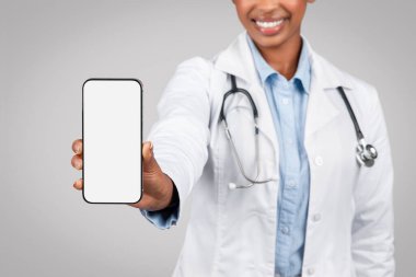 Beyaz önlüklü genç bayan doktorun gri arka plan stüdyosunda izole edilmiş boş ekranlı telefonu göstermesine sevindim. Sağlık hizmetleri, reklam ve sunum sitesi, uygulama tavsiyesi