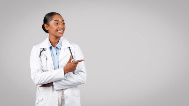 Profesyonel profesyonel siyah bayan doktor, gri arka planda, panorama üzerinde izole edilmiş, boş alanı işaret ediyor. Dikkat dikkat ve teklif, sağlık hizmeti tavsiyesi, sınav