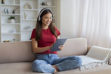 Mutlu Asyalı kadın portresi, evdeki koltukta dijital tabletle dinlenen kablosuz kulaklıklar takan, modern kadın aletlerini ev içi eğlence için kullanan, yeni uygulamaları tarayan,