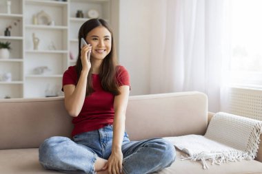Evde cep telefonuyla konuşan gülümseyen Asyalı genç bir kadın oturma odasında koltukta oturup arkadaşlarıyla sohbet ederken mutlu Koreli güzel bir kadının keyifli cep telefonu sohbeti.