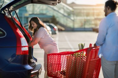 Genç bir kadın, alışverişi başarıyla tamamladıktan sonra açık havada güneşli bir otoparkta alışveriş arabasıyla bekleyen bir adamla birlikte marketin bagajına bir şeyler koyuyor.