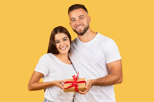 Glad young european guy hug wife, gives box present, enjoy holidays, birthday and New Year, Xmas, isolated on yellow studio background. Gifts to Valentine day, anniversary
