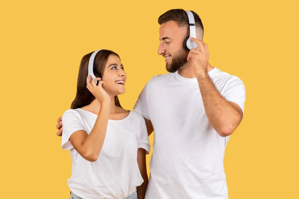 Smiling young european man in white t-shirt and lady in wireless headphones, listen music, have fun, isolated on yellow studio background. Spare time, online audio app, dance