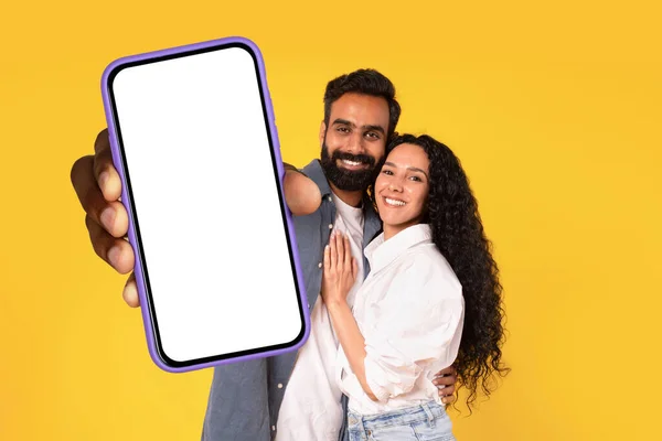 stock image Joyful arabic spouses hug and show large cellphone with blank touchscreen, advertising applications and modern gadget concepts. studio shot on yellow background, collage