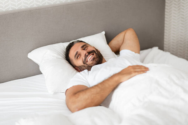 Доброе утро. Счастливый парень, проснувшись, лежит в удобной кровати, прикрытый одеялом в современном помещении, держа голову в руках. Концепция рекреации и отдыха