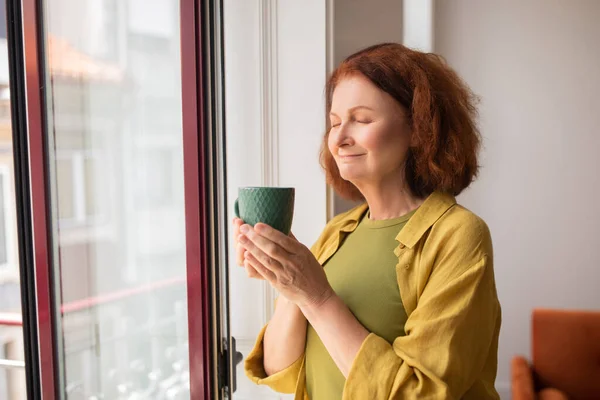 Peaceful senior woman inhaling hot tea aroma while standing near window at home, smiling elderly lady with eyes closed relaxing in domestic interior, enjoying quiet moment in her daily life