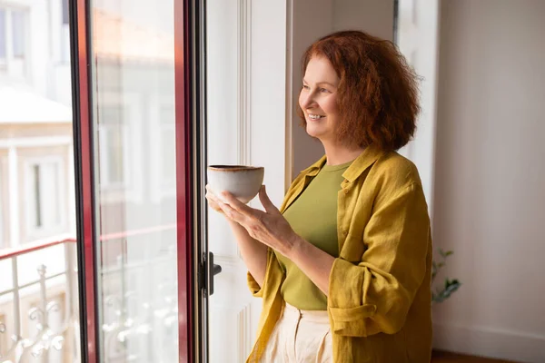 自宅の窓の近くに立っている間 笑顔のシニア女性はコーヒーを飲みながら 街並みを眺めながらリラックスした高齢女性 落ち着いた国内朝と退職時間 空きスペース — ストック写真