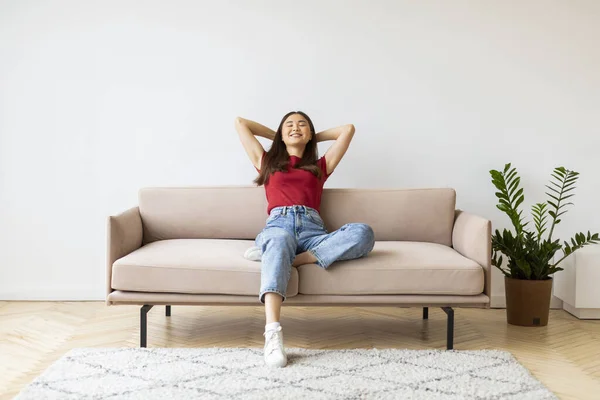 Home Comfort. Portrait Of Smiling Young Asian Woman Leaning Back On Couch, Happy Millennial Lady Resting On Sofa In Living Room Interior, Relaxing With Closed Eyes And Hands Behind Back, Copy Space