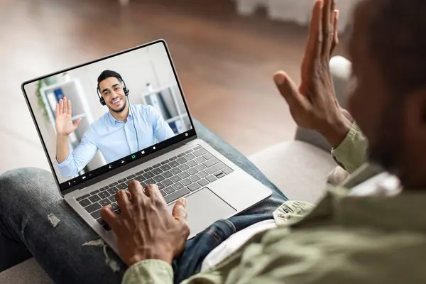 Happy adult black man using computer, have video call with middle eastern guy customer service representative, waving hand at webcam, sit on sofa in living room interior
