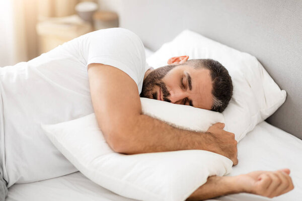 Мужчина средних лет спит дома, лежа в уютной кровати в помещении. Бородатый парень мирно обнимает подушку, быстро спит с закрытыми глазами в современном интерьере спальни. Концепция отдыха