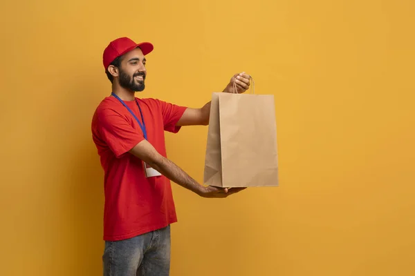Cheerful delivery man in red uniform and cap extending brown paper bag with order, friendly smiling courier guy standing on yellow studio background, recommending fast delivery services, copy space