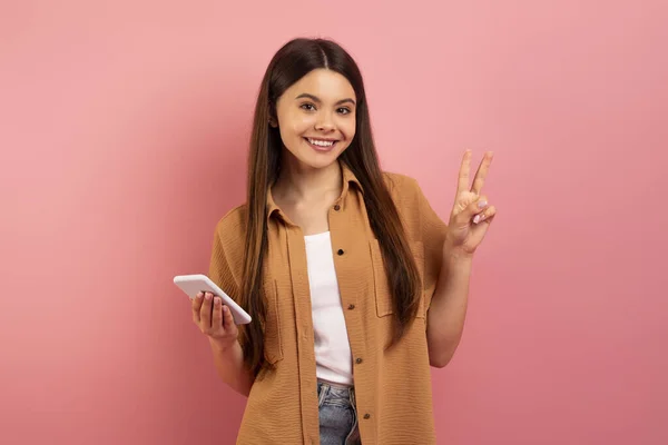 快乐的少女拿着智能手机 做着和平手势 快乐的少女用手机 展示V字形 年轻女子站在粉红工作室的背景上 复制空间 — 图库照片