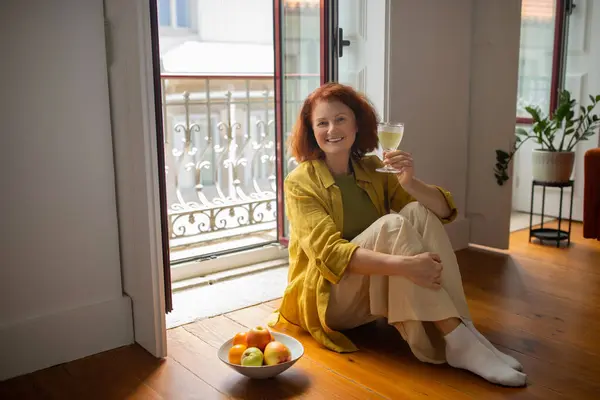 白ワインのグラスと近くの新鮮なフルーツのボウルで床に座っている笑顔のシニア女性 オープンバルコニーのドアで居心地の良い家の雰囲気でリラックスした幸せな高齢女性 退職時間を楽しむ — ストック写真