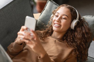 Dijital Eğlence. Kablosuz kulaklıklı genç kız akıllı telefon kullanıyor, müzik dinliyor ve sosyal medyada mesajlaşıyor, evdeki koltukta yatıyor. Gençlik ve Gadgets Eğlence Konsepti