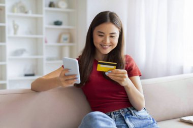 E-Ticaret Konsepti. Genç Asyalı kadın akıllı telefon ve kredi kartı kullanıyor evde koltukta otururken, gülümseyen Koreli kadın internet üzerinden ödemelerini yapıyor cep telefonunda uygulaması, kopyalama uzayı