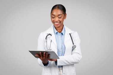 Beyaz önlüklü gülümseyen genç siyahi kadın doktor tablette yazı yazıyor, cihazda çalışıyor, gri arka plan stüdyosunda izole edilmiş. Modern tıp, destek uygulaması, online muayene hastası