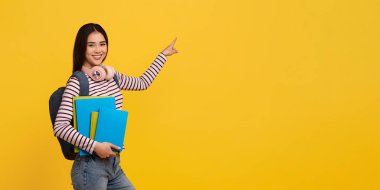 Sırt çantalı, kablosuz kulaklıklı ve elinde reklam için fotokopi alanını gösteren kitapları olan neşeli Asyalı genç bayan öğrenci, sarı stüdyo arka planı, web-afişi. Eğitim