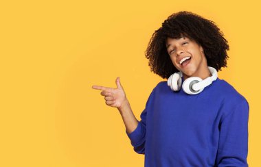 Boynunda kablosuz kulaklıkları olan neşeli genç siyah adam fotokopi alanını işaret ediyor. Sarı arka planda reklam için bedava yer gösteren mutlu bir Afrikalı Amerikalı adam.