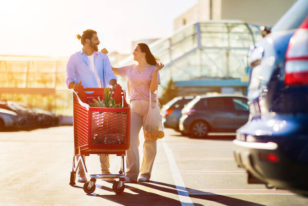 Happy arab man and latin woman with full grocery cart walking together in a sunny parking lot, having a cheerful conversation, full length