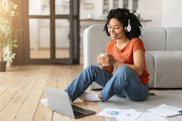 Smiling Black Woman In headphones Using Laptop And Drinking Coffee At Home, Happy African American Freelancer Lady Sitting On Floor In Living Room, Working With Computer And Enjoying Hot Drink