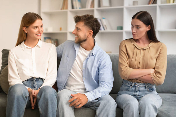 Young man hugs his girlfriend on couch while another woman looks with jealousy crossing hands, sitting together on sofa in modern living room. Infidelity, complicated romantic triangle