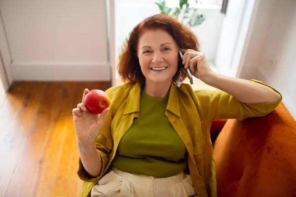ハッピーレッドヘッドシニア女性は 携帯電話で話し 自宅でAppleを食べる 携帯電話の会話を持っている陽気な老婦人とリビングルームでカウチでリラックスしながら健康的なフルーツスナックを楽しむ — ストック写真